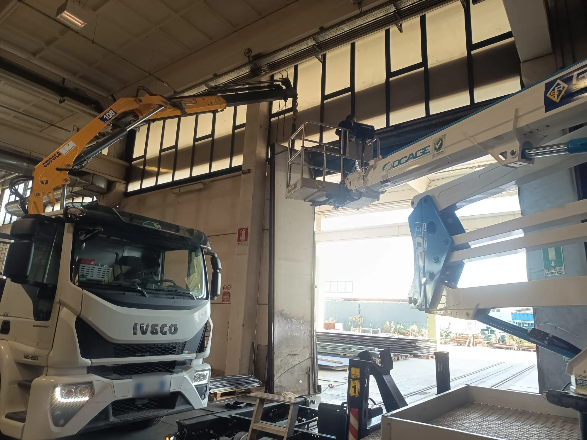 Autocarro con gru dentro un capannone, accanto a una piattaforma aerea con un operaio al lavoro in quota