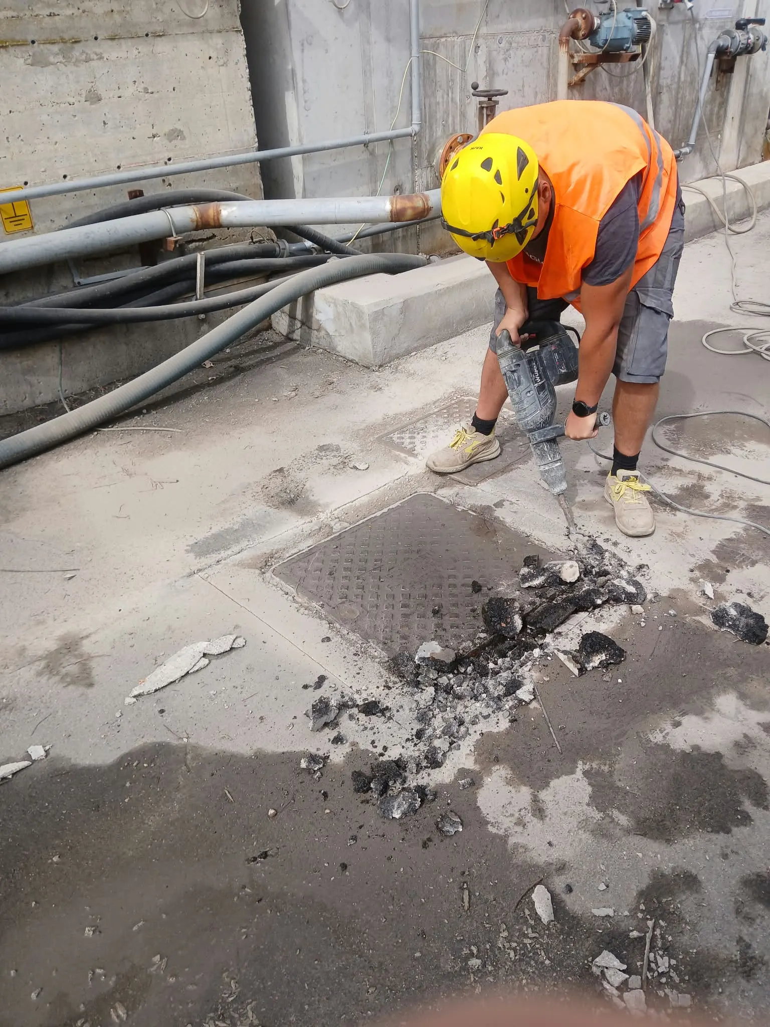 Operaio con casco e martello demolitore che rompe il cemento attorno a un chiusino in uno stabilimento