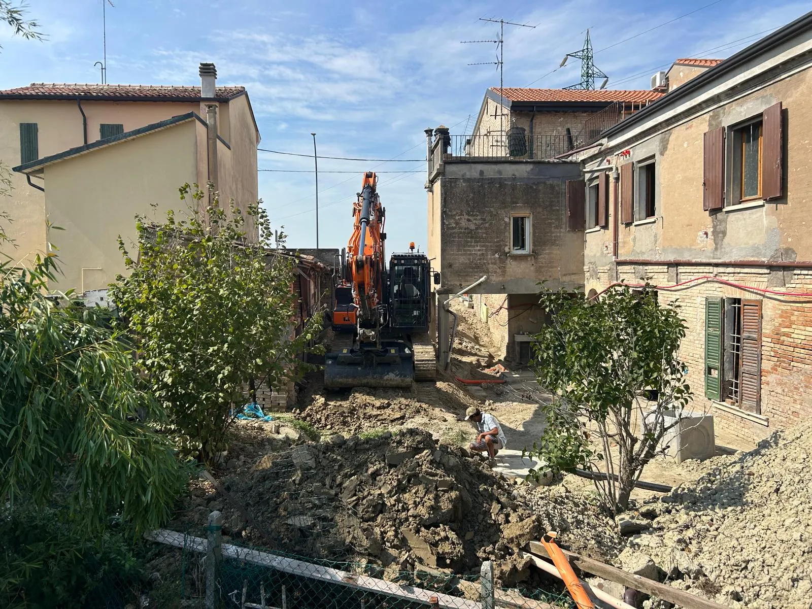 Escavatore che lavora in uno spazio stretto tra due case, con tubi e pozzetti in posa