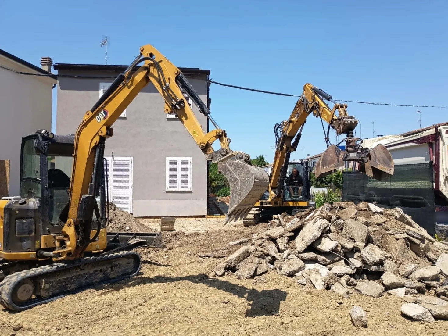 Miniescavatore ed escavatore con pinza che spostano un cumulo di macerie di calcestruzzo davanti a una casa