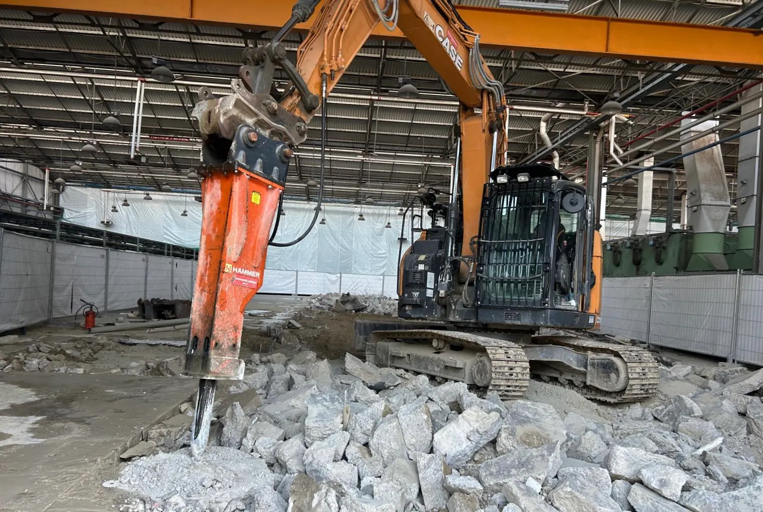 Escavatore con martello idraulico che rompe una pavimentazione in calcestruzzo dentro un capannone industriale