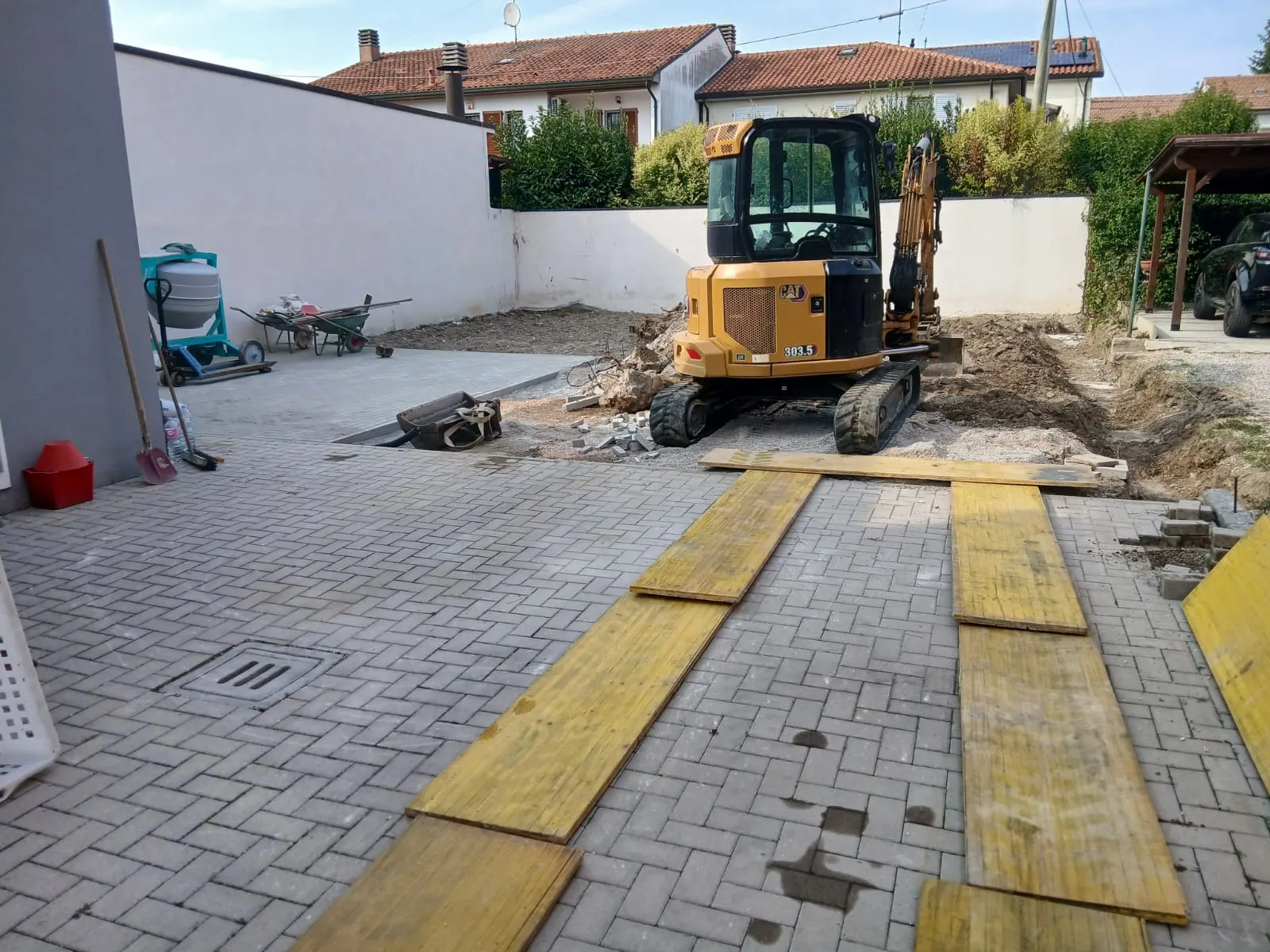 Cortile con un miniescavatore sullo sfondo e, in primo piano, tavole di legno appoggiate sugli autobloccanti per proteggere la pavimentazione esistente