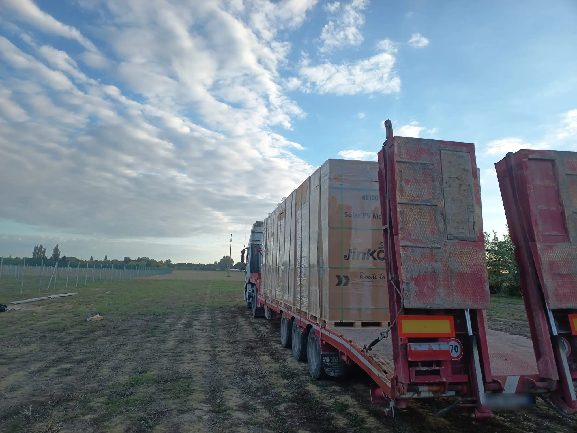 Bilico con pianale ribassato carico di pallet di pannelli fotovoltaici, fermo in un campo accanto al recinto di un impianto
