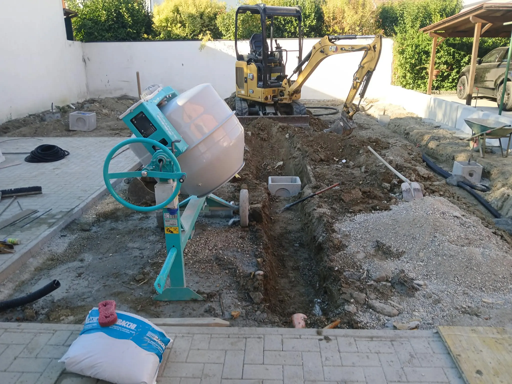 Miniescavatore che scava una trincea nel cortile di una casa, con pozzetti già posati, tubi corrugati e una betoniera